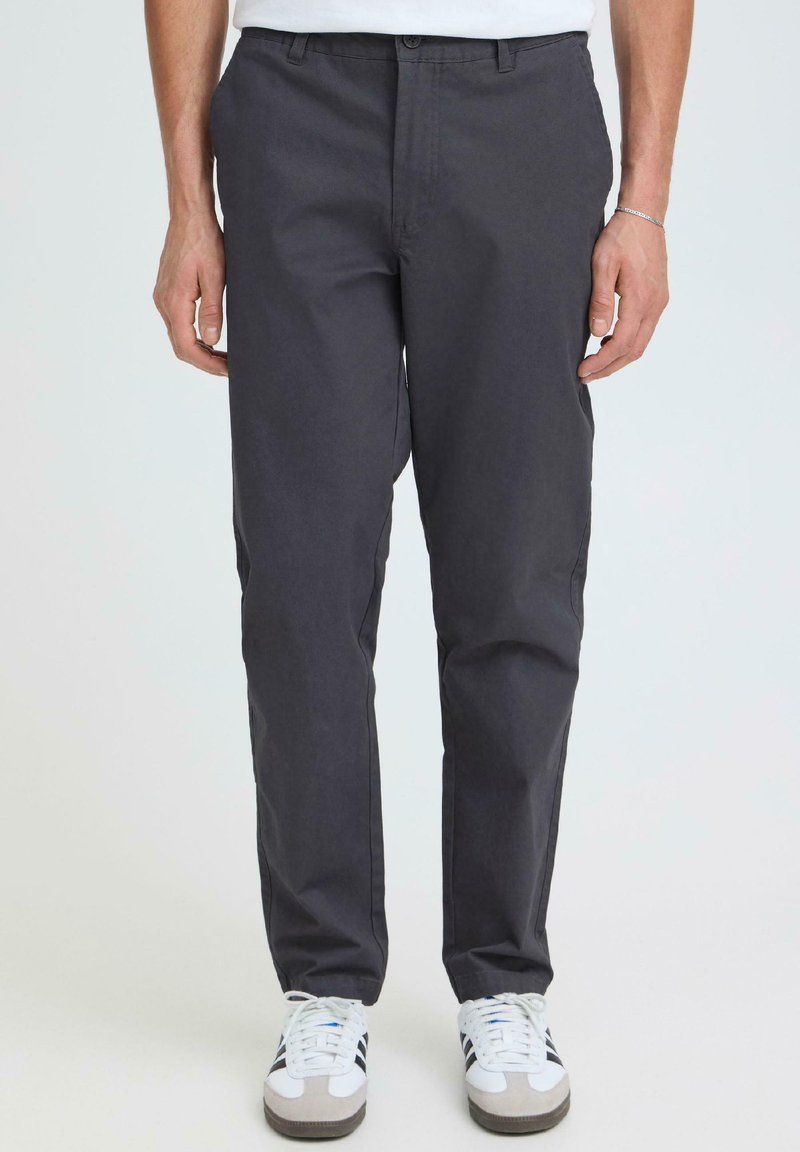 Homme portant un pantalon droit gris foncé et des baskets blanches à rayures noires, debout devant un fond clair uni.