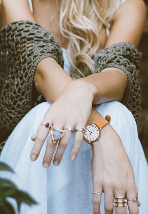 Montre en or rose avec un bracelet orange, plusieurs bagues dont une bague avec une pierre triangulaire, portée sur une peau bronzée, avec un fond de robe blanche.