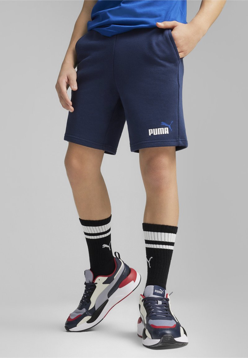 Shorts en coton bleu marine avec poches latérales, arborant un logo Puma blanc. Associés à des chaussettes à rayures noir et blanc et à des baskets multicolores.