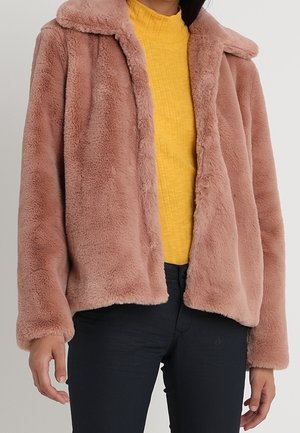 Mujer con una chaqueta de piel sintética marrón claro y suave sobre un jersey amarillo texturizado y pantalones negros sobre un fondo liso.