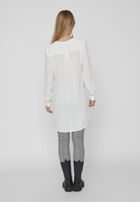 Lång vit blus med ryggbärs, slät struktur och knappade manschetter, tillsammans med rutiga byxor och knähöga svarta stövlar.