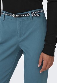 Mēteļi no gludas auduma, ar melnbaltu svītrainu jostu un sudraba elementiem, tuvplāns par kabatu un jostas daļu.