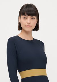 Maillot de bain à manches longues de couleur marine avec une bande horizontale dorée à la taille. Texture lisse avec un design ajusté et un col rond.