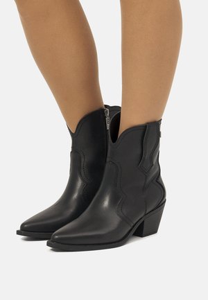 Botas western de cuero negro hasta el tobillo con puntas puntiagudas, cremalleras laterales y tacones bajos apilados, usadas sobre piernas desnudas.