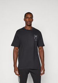 Schwarzes Baumwoll-T-Shirt mit kurzen Ärmeln, das ein Grafikdesign auf der linken Brust zeigt. Modell kombiniert mit dunklen Hosen und steht vor einem einfarbigen Hintergrund.