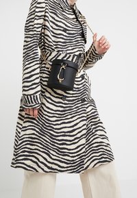 Schwarzer und cremefarbener, zebra gestreifter Trenchcoat mit gebundenem Taillenbund und Knöpfen, kombiniert mit einer kleinen schwarzen zylindrischen Tasche mit einer goldenen Kette und Verschluss.