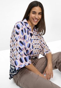 Donna sorridente con lunghi capelli scuri seduta, che indossa una blusa a fantasia geometrica nei toni del rosa, blu e beige e pantaloni marrone chiaro.