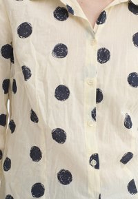 Chemise blanche ornée de grands cercles bleu marine, en tissu léger, avec une fermeture à boutons et une coupe décontractée. Apparence texturée.