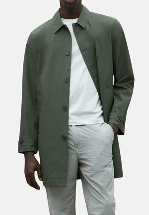 Abrigo verde oliva con corte recto, con cierre de botones y bolsillos laterales, llevado sobre una camisa blanca y pantalones gris claro.