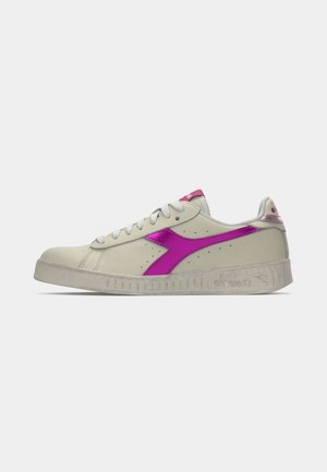 Zapatilla baja color blanco roto con cordones blancos, suela gruesa y acentos metálicos en rosa brillante en el logo y el talón, sobre un fondo liso.