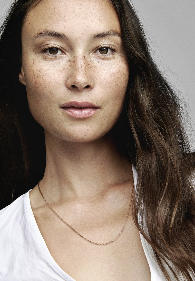 Donna con lunghi capelli marroni mossi e lentiggini, che indossa una maglietta bianca con scollo a V e una semplice collana d'oro. Sfondo neutro.