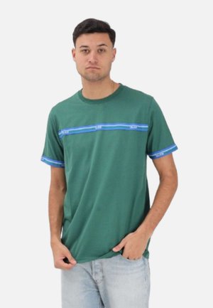 Giovane uomo che indossa una maglietta verde a maniche corte con strisce blu sul petto e sulle maniche, abbinata a jeans azzurri chiari, in piedi su sfondo bianco.