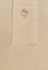 Tissu beige à texture lisse avec une fermeture à bouton. Comprend une poche plaquée avec des détails de surpiqûres ton sur ton.