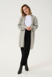 Grau Cardigan aus weichem, strukturiertem Material, offen vorne, mit langen Ärmeln, über einem weißen T-Shirt getragen, kombiniert mit schwarzen Hosen und Ankle Boots.