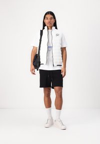 Balta siuvinėta liemenė su juodais akcentais, dėvima ant baltos marškinėlių su mėlynai tekstu, derinama su juodomis trumpomis kelnėmis ir baltomis sportbačiais. Juoda rankinė ant peties.