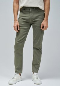 Pantalones ajustados de color verde oliva, hechos de una mezcla de algodón, con bolsillos frontales, cremallera y detalles de costura sutiles. Combinados con zapatillas blancas.