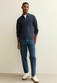 Pullover blu navy con zip, colletto e tasca sul petto, abbinato a jeans blu e sneakers bianche con dettagli marroni.