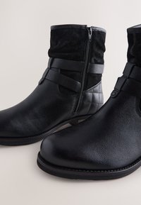 Black ankle boots featuring a leather body, suede upper, side zipper closure, and cross straps. Textured finish and flat sole.