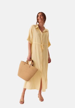 Donna sorridente che indossa un vestito midi giallo a righe con bottoni e cintura in vita, tiene una borsa intrecciata e indossa sandali con tacco color nude.