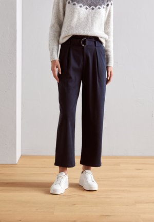 Persona con pantalones cortos de pierna ancha de color azul marino y cinturón, un suéter blanco con estampado y zapatillas blancas con cordones, de pie sobre un suelo de madera.