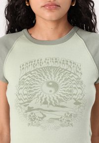 Green cotton t-shirt with short sleeves, featuring a faded, printed sun and yin-yang design at the center, and floral accents at the bottom.