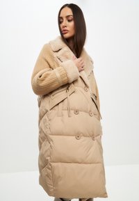 Beige todelt jakke med to materialer, faux fur-krave, kviltet krop og strikkede ærmer. Har sidelukning med lynlås og knapdetaljer.