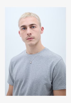 Jaunas žmogus su trumpais blondiniais plaukais, dėvi pilką marškinėlį ir sidabrinį saulės formos pakabuką ant kaklo, žvelgiantis tiesiai į priekį su neutralia veido išraiška.