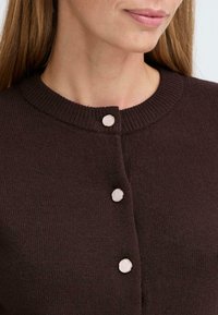 Brauner Strickcardigan mit rundem Halsausschnitt, der an der Vorderseite drei hellrosa Knopfverschlussmöglichkeiten und gerippte Bündchen hat.