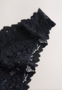 Tissu en dentelle noire avec motifs floraux et bords festonnés posé à plat sur une surface claire, montrant des détails textiles complexes.