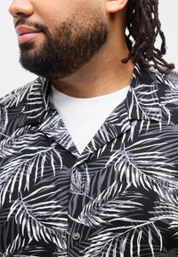 Άνδρας με γένια και κότσους φοράει μαύρο πουκάμισο με κουμπιά, με λευκό και γκρι σχέδιο από φύλλα φοίνικα, πάνω από λευκό μπλουζάκι.