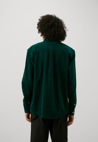 Zielona koszula sztruksowa z długimi rękawami, mankietami na guziki i plisą z tyłu; miękka w dotyku i z zaokrąglonym dołem. Nosi się ją z ciemnymi spodniami cargo.