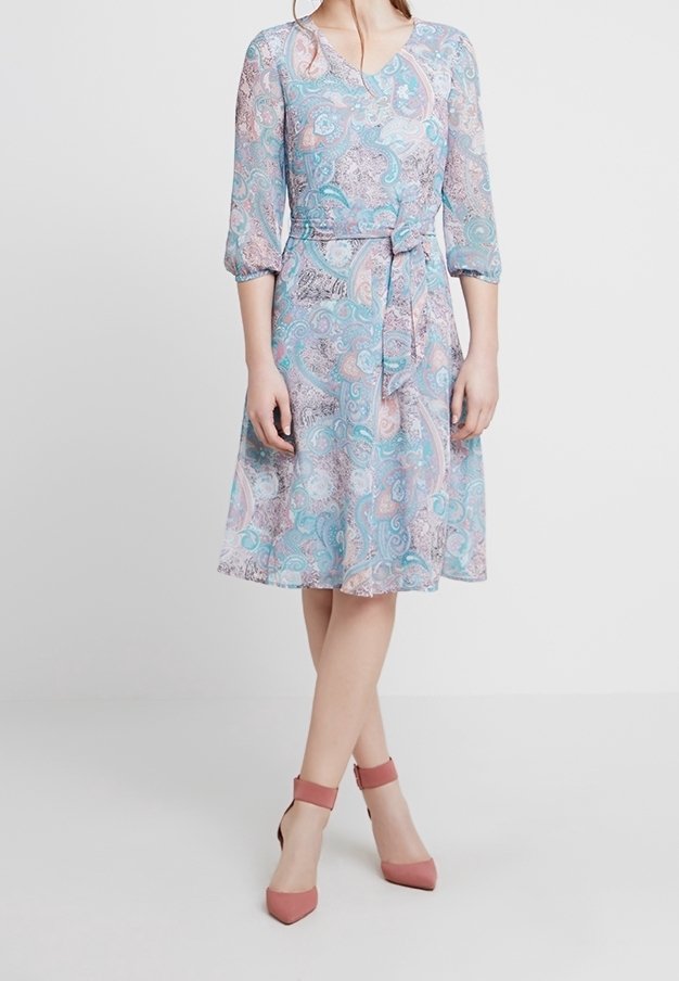 Femme portant une robe portefeuille à motif paisley bleu pastel, rose et blanc, avec des manches trois quarts et des talons à brides mauves, se tenant devant un fond blanc.