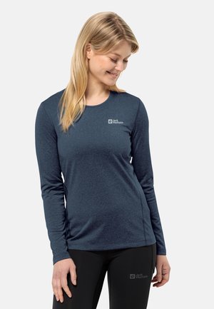 Donna che indossa una maglietta a maniche lunghe blu Jack Wolfskin e leggings neri, sorridente e con lo sguardo rivolto verso il basso a sinistra.