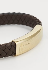 Brunnet vevet lærarmbånd med en gullfarget metalllås som har gravert logo. Designet er strømlinjeformet med en teksturert overflate.