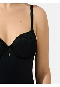 Zwarte lingerie met gepofte cups en verstelbare bandjes. Het materiaal heeft een subtiel geometrisch patroon en heeft een gouden hardware detail in het midden.