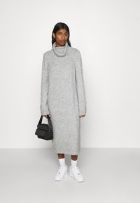 Γκρίζο πλεκτό φόρεμα με ψηλό γιακά και μακριά μανίκια, μήκους μέχρι το γόνατο, υφή υφάσματος, συνδυασμένο με λευκά αθλητικά παπούτσια και μια μικρή μαύρη τσάντα.