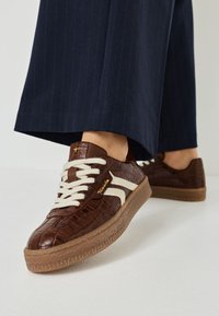 Baskets marron à motif croco avec des accents blancs, dotées de lacets plats crème, d'une semelle en caoutchouc texturée, et d'un logo doré sur le côté.