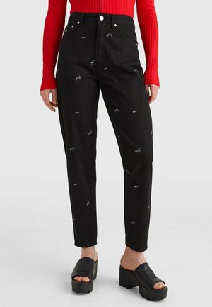 Personne portant un pantalon noir taille haute avec de petits logos blancs, un haut rouge à manches longues et des chaussures noires à plateforme sans lacets.