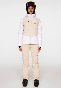 Strój narciarski składający się z białej i beżowej izolowanej kurtki z zamkiem błyskawicznym z przodu, w zestawie z beżowymi spodniami oraz czarnymi butami narciarskimi. Zawiera kask i gogle.