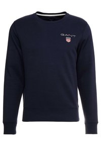 Sudadera de color azul marino con mangas largas, cuello redondo y puños de punto. Presenta un logo de GANT prominente y un diseño de escudo. Tela suave.
