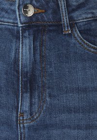 Primer plano de unos jeans de denim azul que muestra el bolsillo frontal, el lazo del cinturón, las costuras naranjas y el botón de metal con la grabación de la marca.