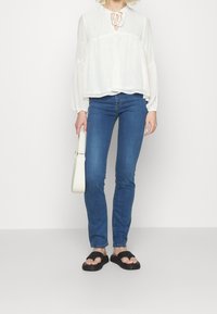 Blusa blanca de manga larga con cuello redondo y detalle de lazo, combinada con jeans ajustados azules y sandalias planas negras. Sosteniendo un bolso pequeño blanco.