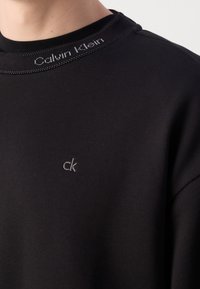 Gros plan d'une chemise noire Calvin Klein montrant le col avec le texte "Calvin Klein" et un petit logo "ck" sur la poitrine.
