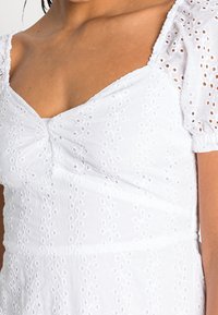 Vitt bomullsklänning med puffärmar, fyrkantig ringning och flätad broderi med en rynkad bystdetalj.