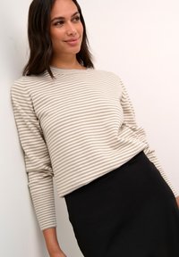 Pull rayé beige et gris, avec des manches longues et un col arrondi, associé à une jupe noire. Texture lisse, coupe décontractée.