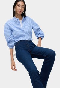 Kobieta w jasnoniebieskiej koszuli w paski z guzikami i ciemnoniebieskich dżinsach, siedząca z jednym ramieniem opartym na kolanie na białym tle.