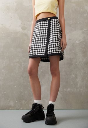 Femme portant une mini-jupe pied-de-poule noire et blanche avec bordure noire, un top court jaune sans manches, des chaussettes blanches et des bottes noires épaisses.