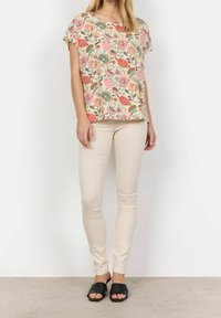 Blumenbluse mit kurzen Ärmeln, verziert mit rosa, orange und grünen Mustern. Kombiniert mit beigen Skinny-Hosen und schwarzen Rutschsandalen.