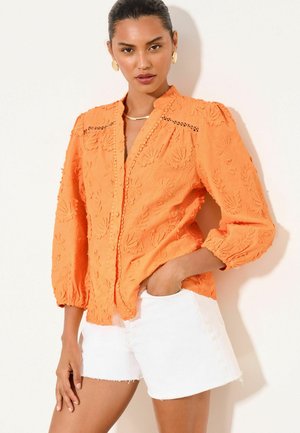 Frau trägt eine orange bestickte Bluse mit Puffärmeln und weißen Shorts, steht mit einer Hand in der Tasche vor neutralem Hintergrund.