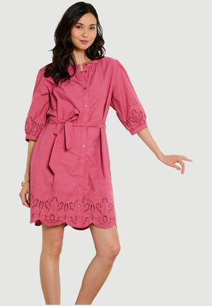 Frau trägt ein knielanges rosa Kleid mit durchgehender Knopfleiste, gebundener Taille, Puffärmeln und welligem Saum mit bestickten Ausschnittdetails.